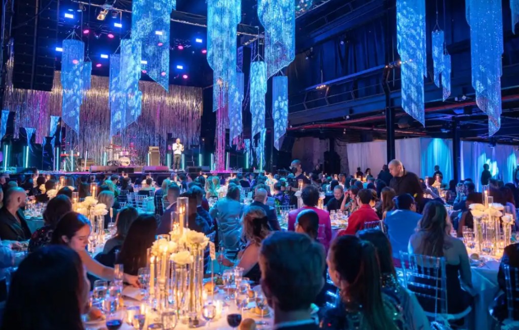 A corporate gala dinner with hanging blue lighting, candlelit tables, and a stage beyond the room.